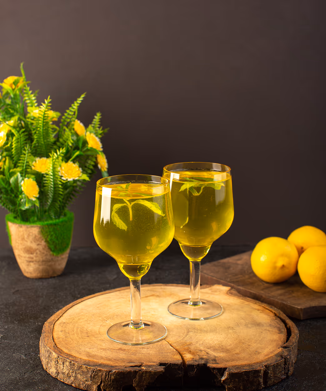 Two glasses of yellow drink garnished with mint leaves on a wooden slice with lemons and a yellow flower plant in the background.